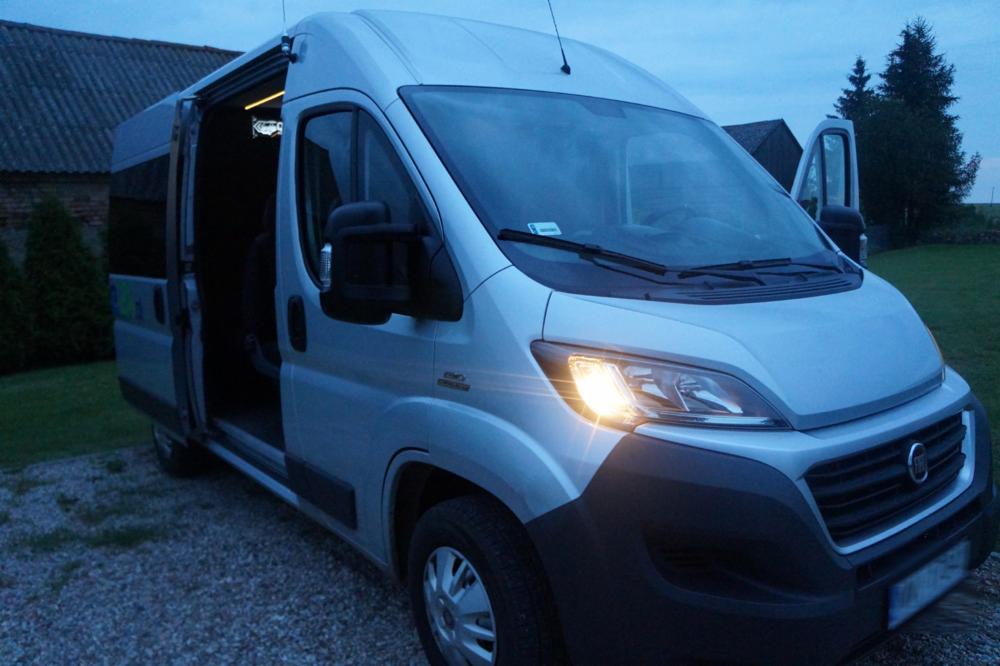 Srebrny van z otwartymi drzwiami bocznymi stoi na żwirowym podłożu przed budynkiem z ciemną dachówką, z włączonym reflektorem.