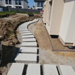 Ścieżka z jasnych, prostokątnych płyt prowadząca przez ogród wzdłuż domu. Widoczne przygotowane podłoże i fragmenty trawnika. Nowoczesne budynki w tle.