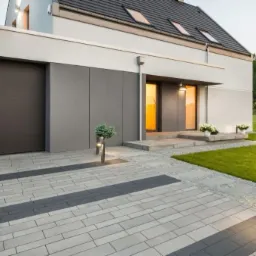 Nowoczesny dom z szarą kostką brukową ułożoną we wzór. Brama wjazdowa i garaż w kolorze antracytowym. Oświetlenie zewnętrzne.