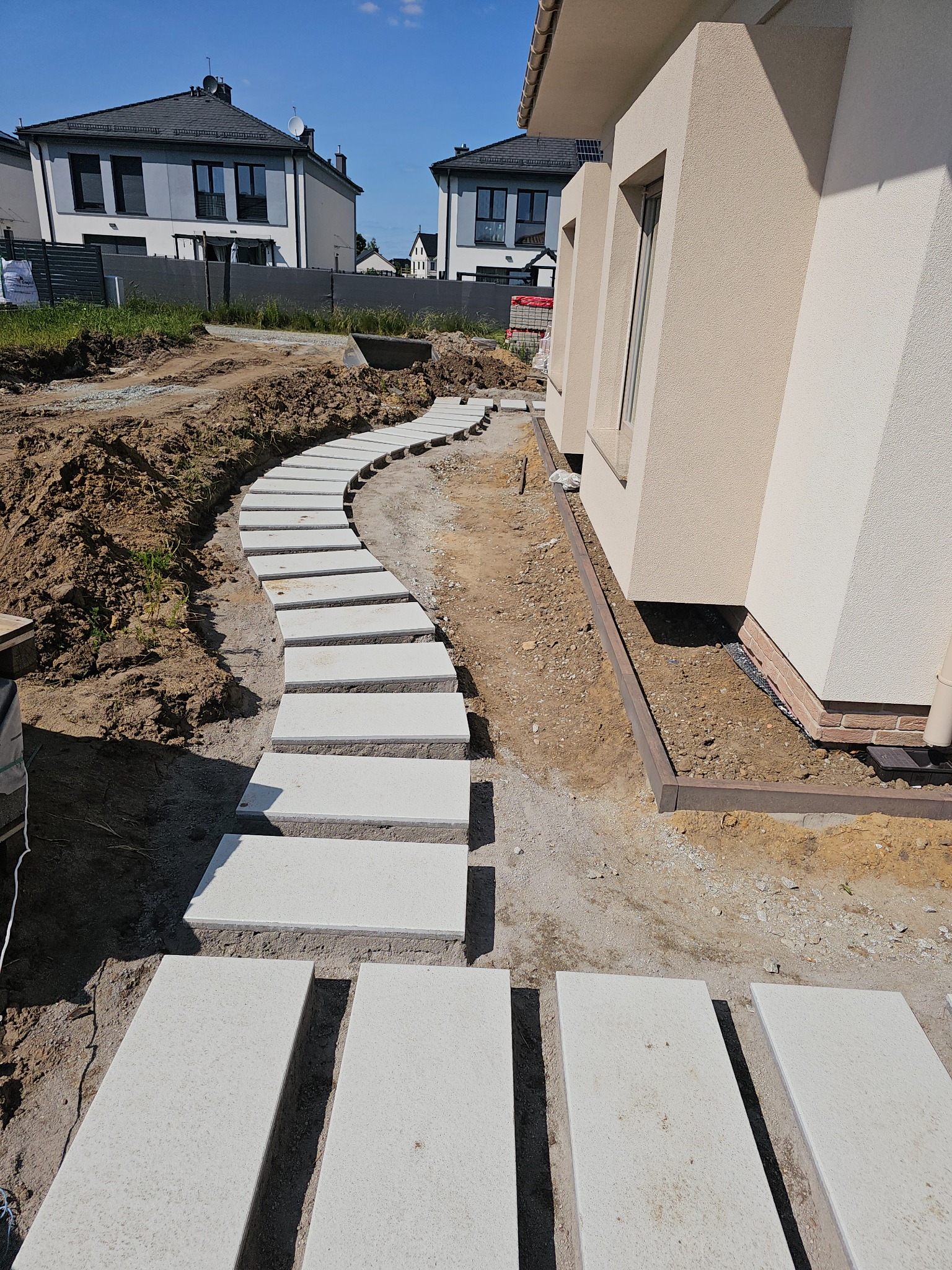 Ścieżka z jasnych, prostokątnych płyt prowadząca przez ogród wzdłuż domu. Widoczne przygotowane podłoże i fragmenty trawnika. Nowoczesne budynki w tle.