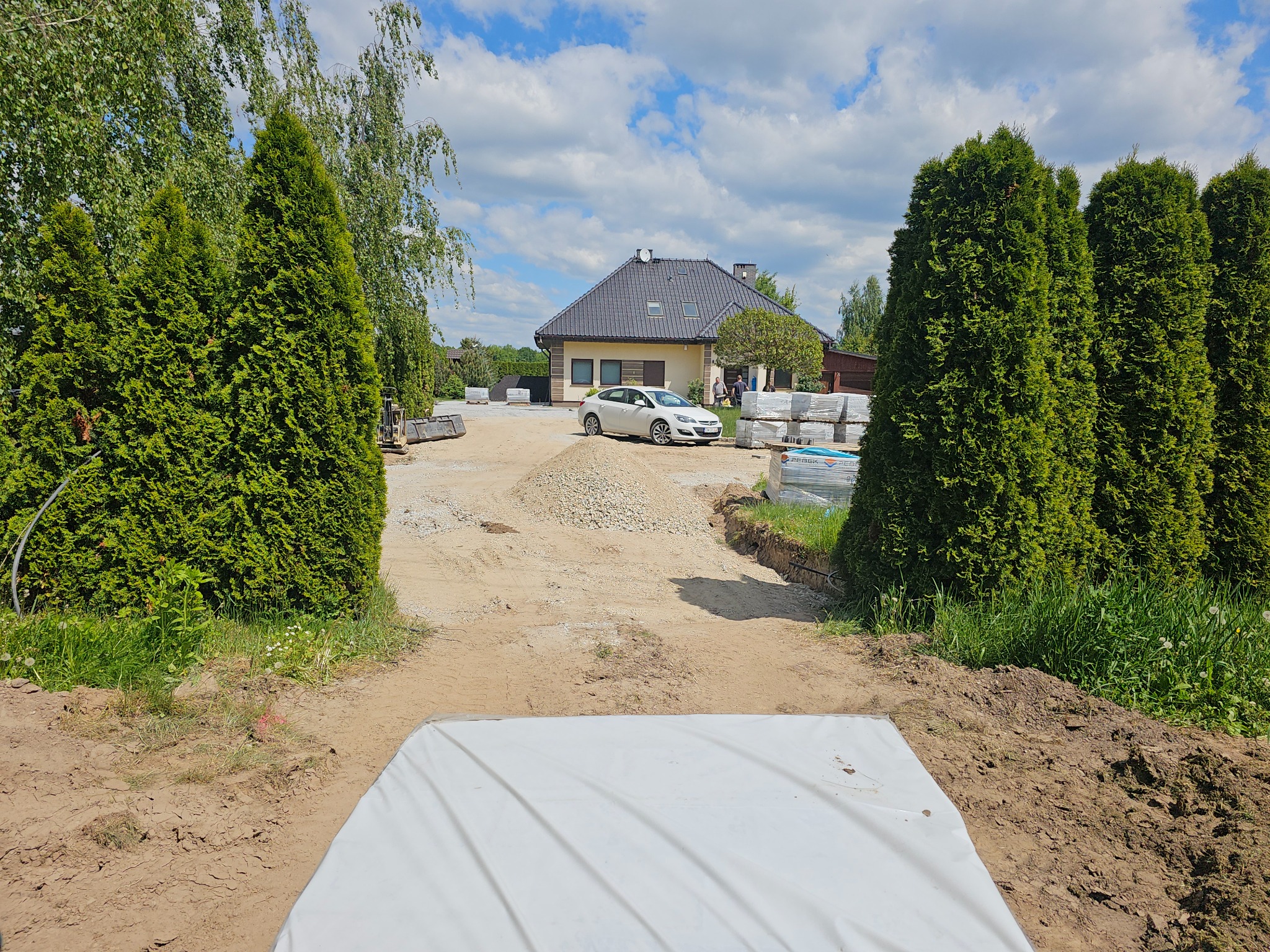 Przygotowanie podłoża pod kostkę: wyrównany teren z podsypką, obrzeża wykopane, w tle dom jednorodzinny i zaparkowany samochód, słoneczny dzień.