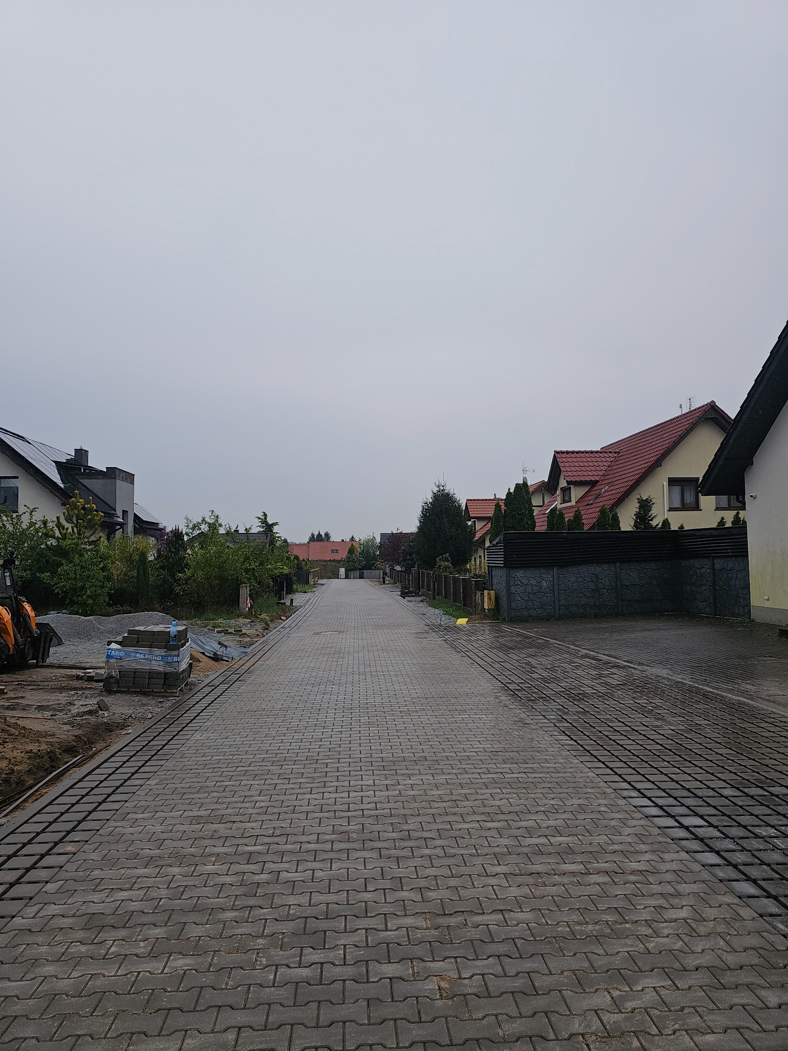 Długa droga z szarej kostki brukowej między domami, częściowo ułożona, z widocznymi paletami kostki i mini koparką. Pochmurne niebo.