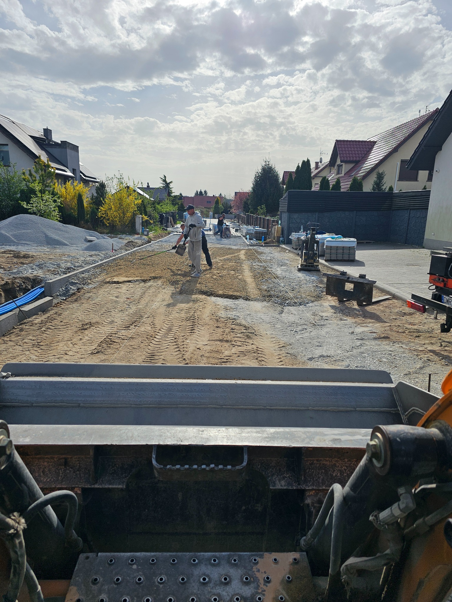 Prace brukarskie na osiedlowej drodze: robotnicy wyrównują podłoże pod kostkę, widoczne obrzeża, maszyny budowlane i sterty materiałów. Ujęcie z perspektywy maszyny budowlanej.