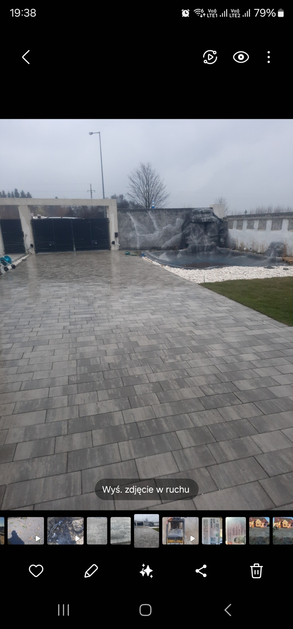 Ułożona, szara kostka brukowa prowadząca do bramy z elementami wodnymi i zielenią. Widok na posesję w pochmurny dzień. Regularny wzór ułożenia kostki.