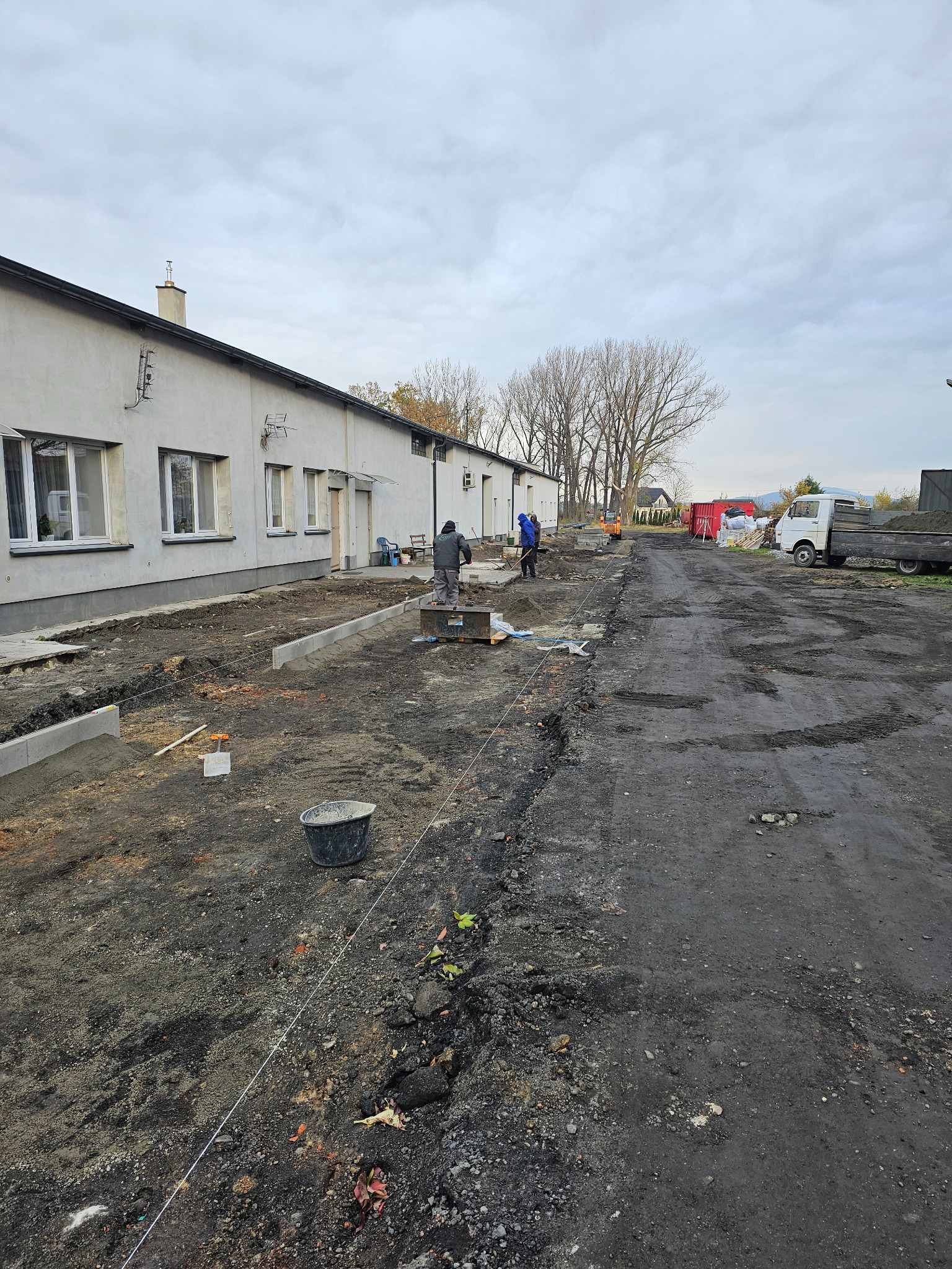 Prace przygotowawcze do ułożenia kostki brukowej. Widoczne obrzeża, pracownicy i sprzęt. Początek realizacji przy budynku gospodarczym w pochmurny dzień.