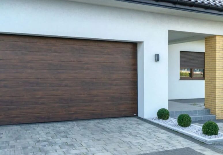 Nowoczesny podjazd z szarej kostki brukowej prowadzący do domu z brązową bramą garażową i białymi ścianami. Dekoracyjne kuliste krzewy wzdłuż podjazdu.