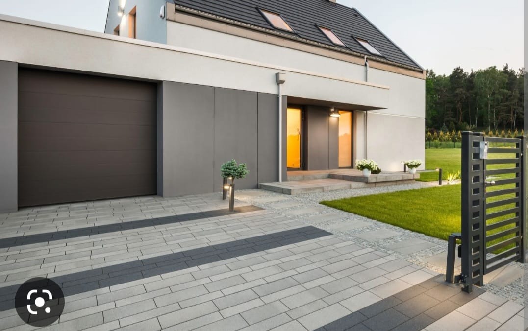 Nowoczesny dom z szarą kostką brukową ułożoną we wzór. Brama wjazdowa i garaż w kolorze antracytowym. Oświetlenie zewnętrzne.