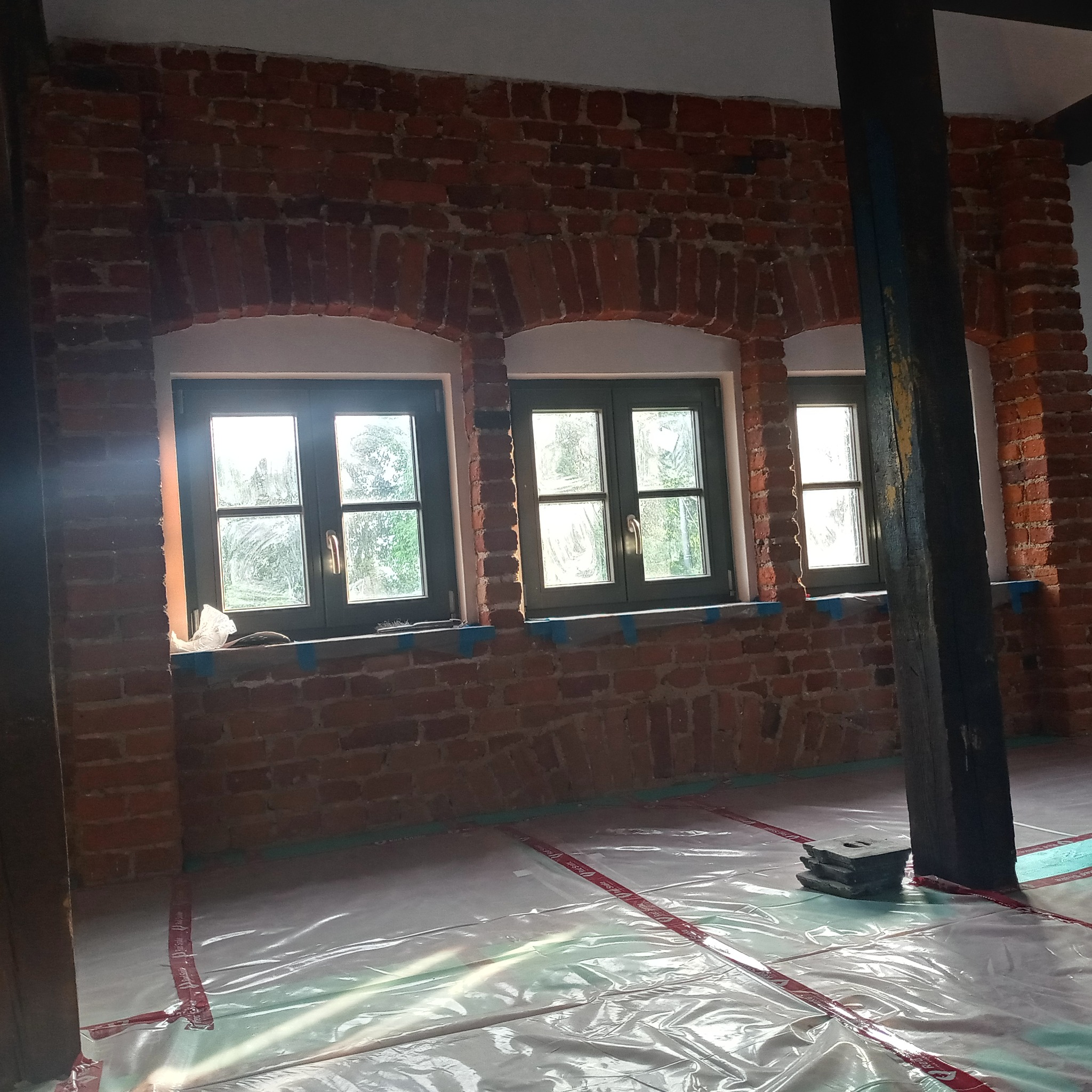 Wnętrze z ceglaną ścianą, trzema oknami z zielonymi ramami i drewnianym słupem, zabezpieczone folią malarską na podłodze, widoczne ślady prac remontowych.