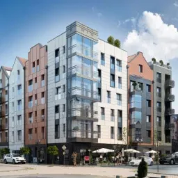 Nowoczesny kompleks budynków mieszkalnych z elementami architektury nawiązującymi do historycznej zabudowy miasta, widok z poziomu ulicy z przechodniami i zaparkowanymi samochodami.