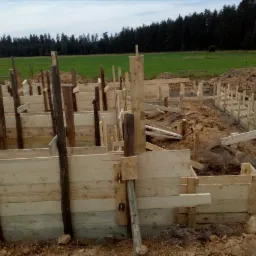 Przygotowanie szalunków pod fundamenty domu jednorodzinnego, widoczne drewniane konstrukcje i ziemia, w tle łąka i las.