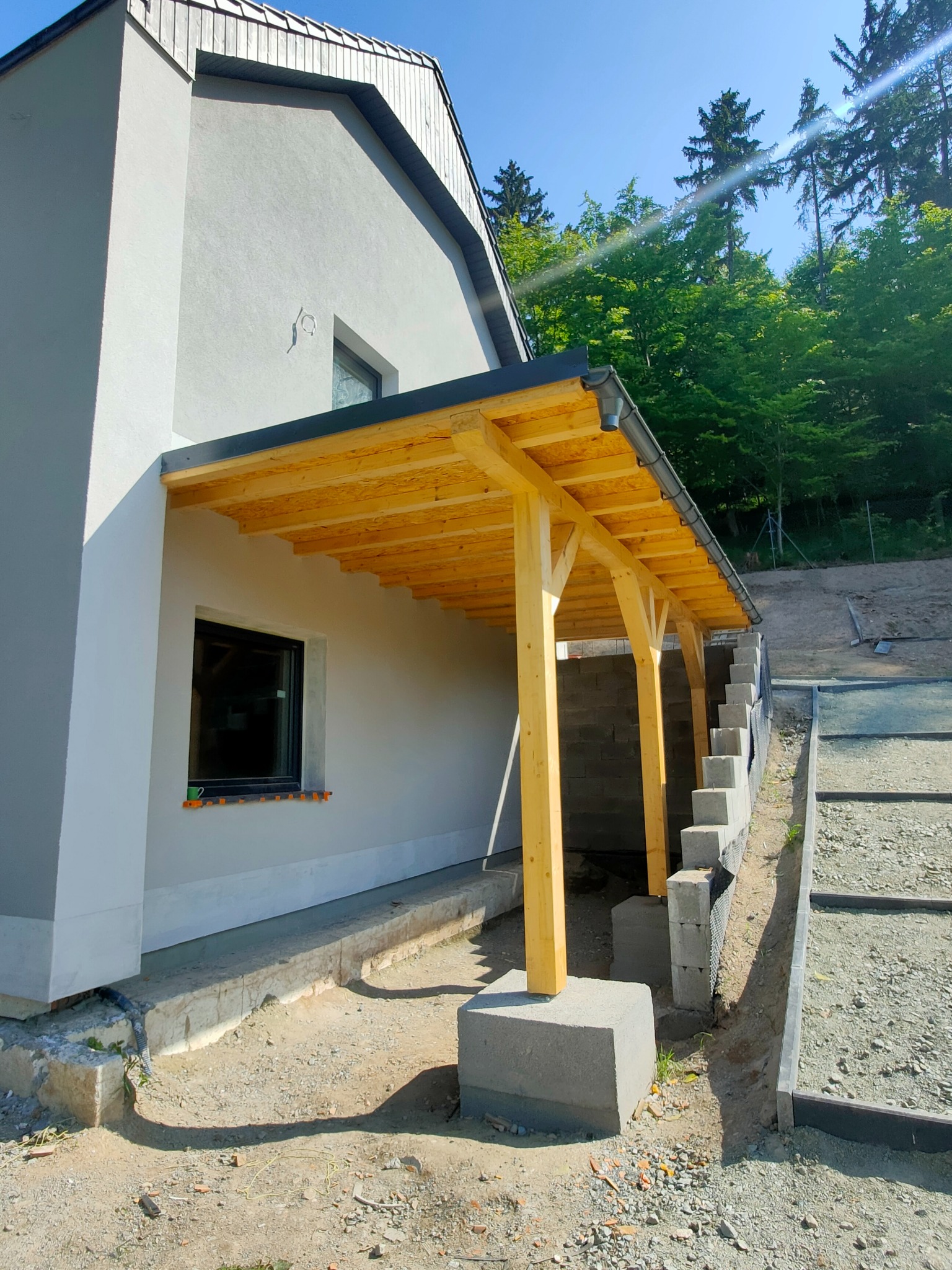 Surowa drewniana konstrukcja zadaszenia przy ścianie domu z widocznym systemem odwadniania, fundamentem i przygotowanym terenem pod dalsze prace.