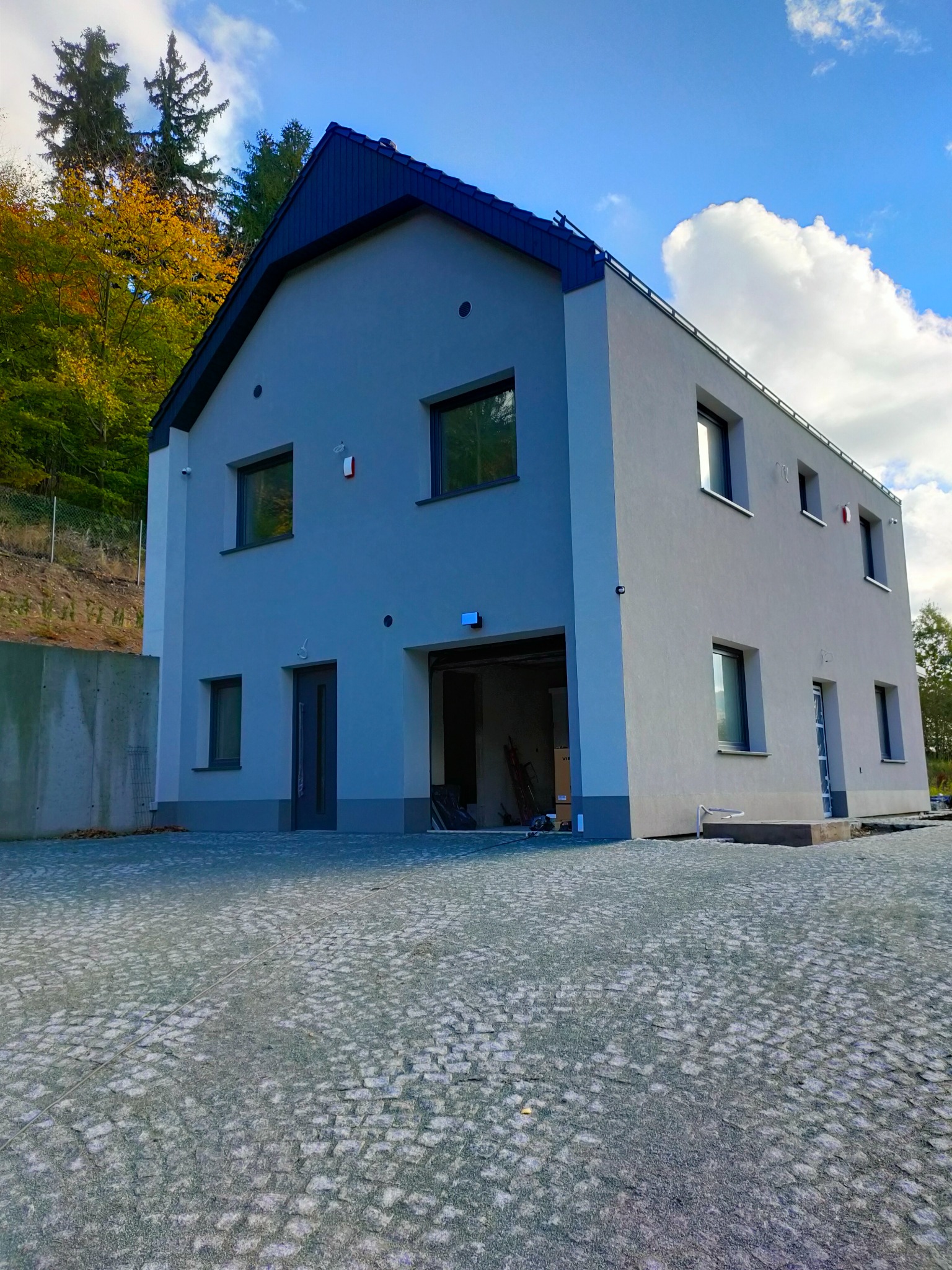 Nowoczesny, dwukondygnacyjny dom z garażem, wykończony na szaro, z ciemnym dachem, widok z zewnątrz na tle zieleni i błękitnego nieba z chmurami.