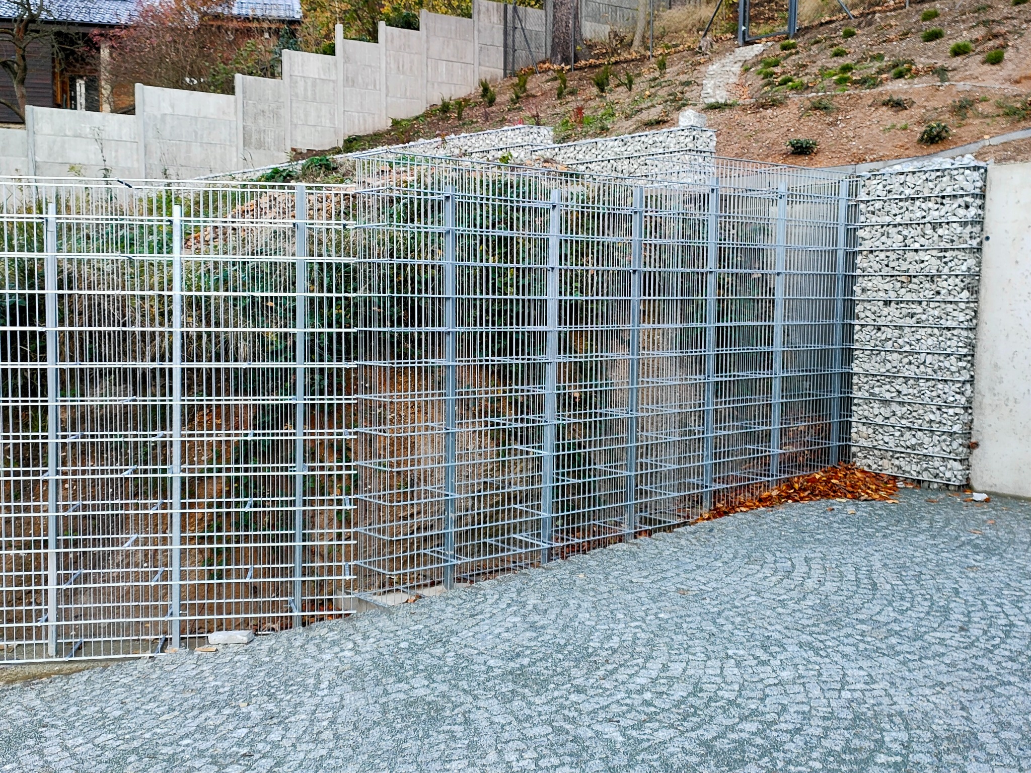Metalowa siatka kaskadowo zabezpieczająca skarpę, wypełniona kamieniami, z widoczną kostką brukową i betonowym murem oporowym w tle.