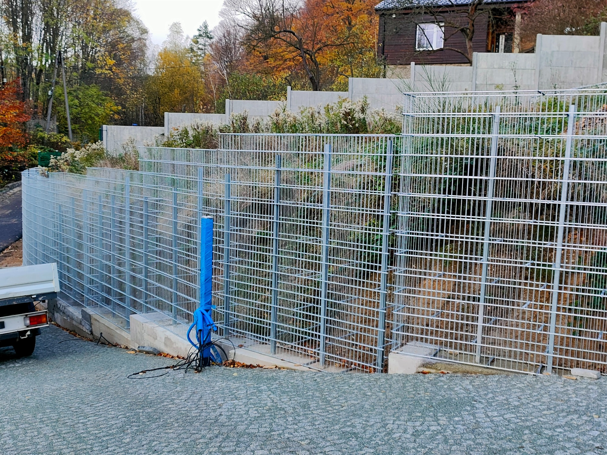 Nowoczesne ogrodzenie panelowe z metalowej siatki na betonowej podmurówce, zabezpieczające skarpę z roślinnością, widok z dołu na tle jesiennego krajobrazu i fragmentu budynku.