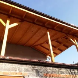 Drewniana konstrukcja zadaszenia tarasu z widocznymi elementami więźby dachowej, brązowe rynny i ceglany mur w tle.