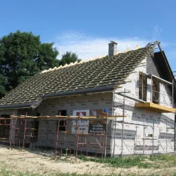 Budowa domu z drewnianą konstrukcją szkieletową w trakcie realizacji, widoczne rusztowania, membrana na ścianach i drewniane łaty na dachu przed położeniem pokrycia.