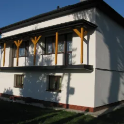 Dwukondygnacyjny dom z białymi ścianami, drewnianą konstrukcją balkonu i ciemnym dachem, widoczny z zewnątrz w słoneczny dzień.