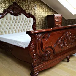 Bogato zdobione, drewniane łóżko w stylu Ludwika z tapicerowanym zagłówkiem w kolorze kremowym, na tle wzorzystej tapety i drewnianej szafki nocnej.