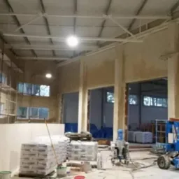 Wnętrze hali w trakcie tynkowania maszynowego; widoczne worki z zaprawą, agregat tynkarski i podnośnik koszowy.