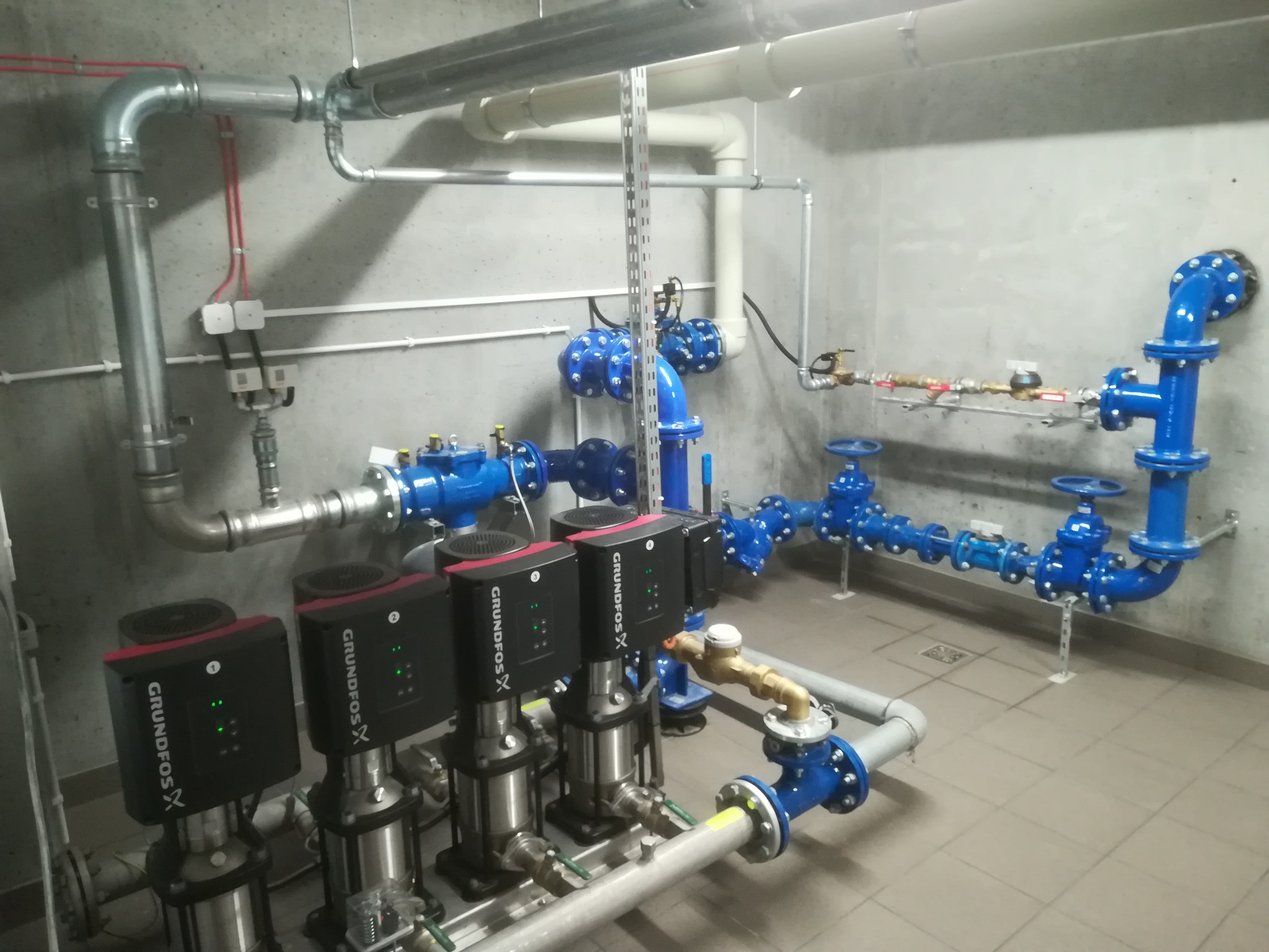Kompleksowa instalacja hydrauliczna z trzema pompami Grundfos X, widoczne rury w kolorze srebrnym, białym i niebieskim, oraz szara ściana i podłoga z płytek.