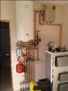 Nowoczesna instalacja grzewcza z białym zasobnikiem ciepłej wody, miedzianymi rurami, rozdzielaczem i kotłem na paliwo stałe w kotłowni.