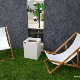 Dwa białe leżaki z drewnianymi ramami i kwadratowa donica na trawie, na tle szarej, teksturowanej ściany i odbijających kul. Jeden leżak złożony, drugi rozłożony.