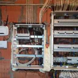 Dwa rozdzielnice elektryczne w metalowych obudowach, zamontowane na surowej ścianie z czerwonej cegły, z widocznymi przewodami i zabezpieczeniami, oraz router WiFi TP-Link i modem D-Link obok.