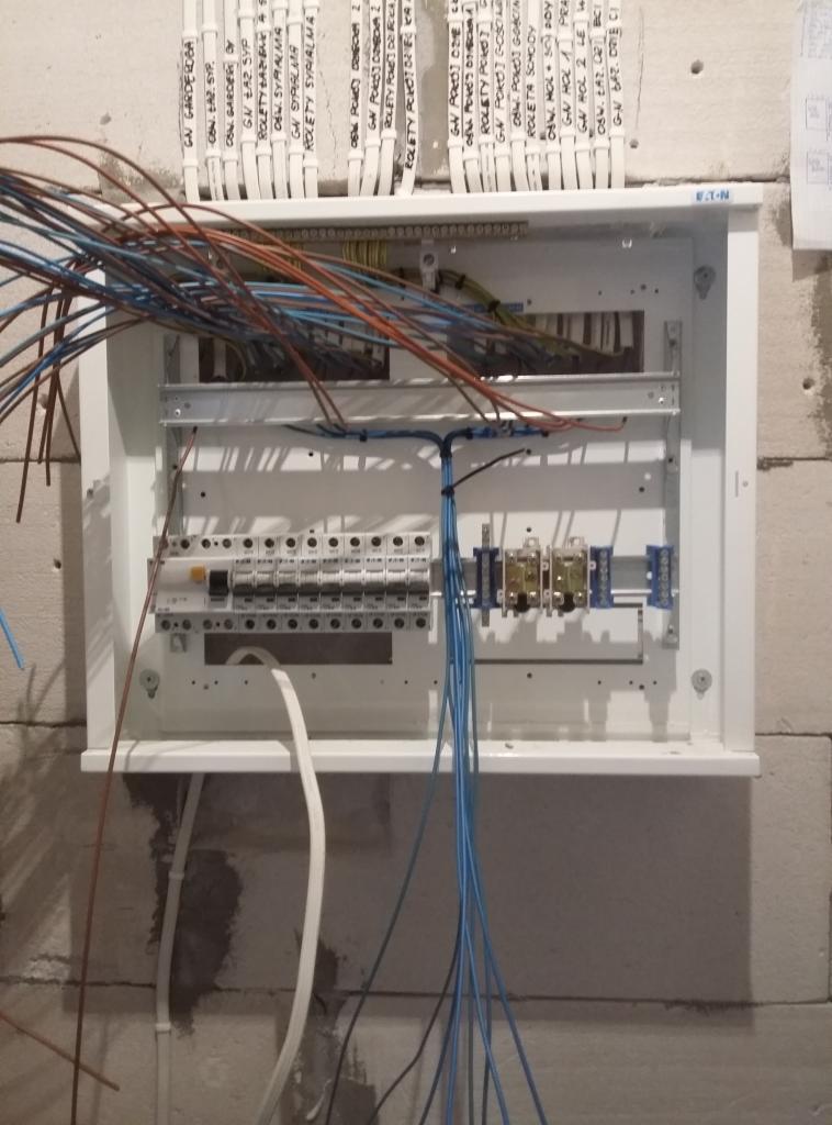 Otwarta skrzynka rozdzielcza Eaton zamontowana na ścianie z betonu komórkowego, widoczne liczne przewody elektryczne w różnych kolorach, etykiety z opisem obwodów nad skrzynką.