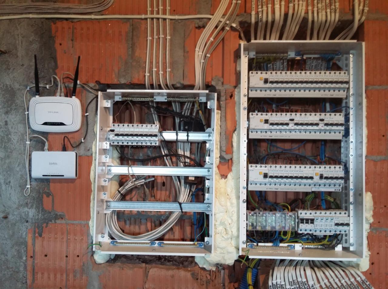 Dwa rozdzielnice elektryczne w metalowych obudowach, zamontowane na surowej ścianie z czerwonej cegły, z widocznymi przewodami i zabezpieczeniami, oraz router WiFi TP-Link i modem D-Link obok.
