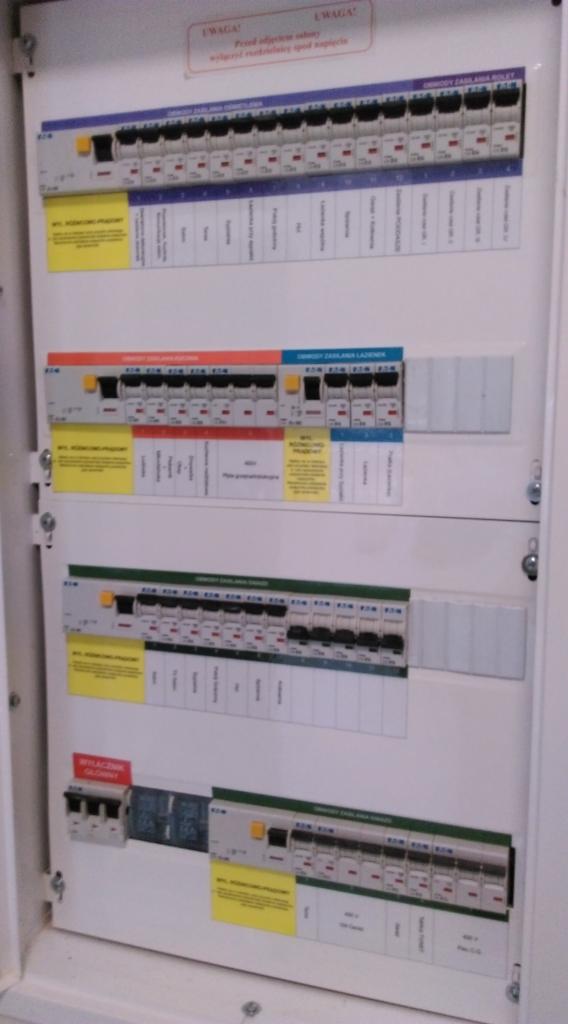 Rozdzielnica elektryczna z bezpiecznikami i naklejkami informacyjnymi, widok z bliska na zabezpieczenia różnicowoprądowe i nadprądowe w skrzynce bezpiecznikowej.