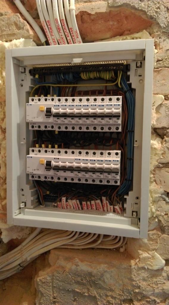 Otwarta skrzynka rozdzielcza z bezpiecznikami Eaton zamontowana na surowej, ceglanej ścianie z widocznymi przewodami i pianką montażową.