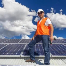 Instalator w kasku i kamizelce odblaskowej stoi na dachu pokrytym panelami słonecznymi, trzymając wiertarkę na tle błękitnego nieba z obłokami; widoczne logo firmy 'DOM-GAZ'.