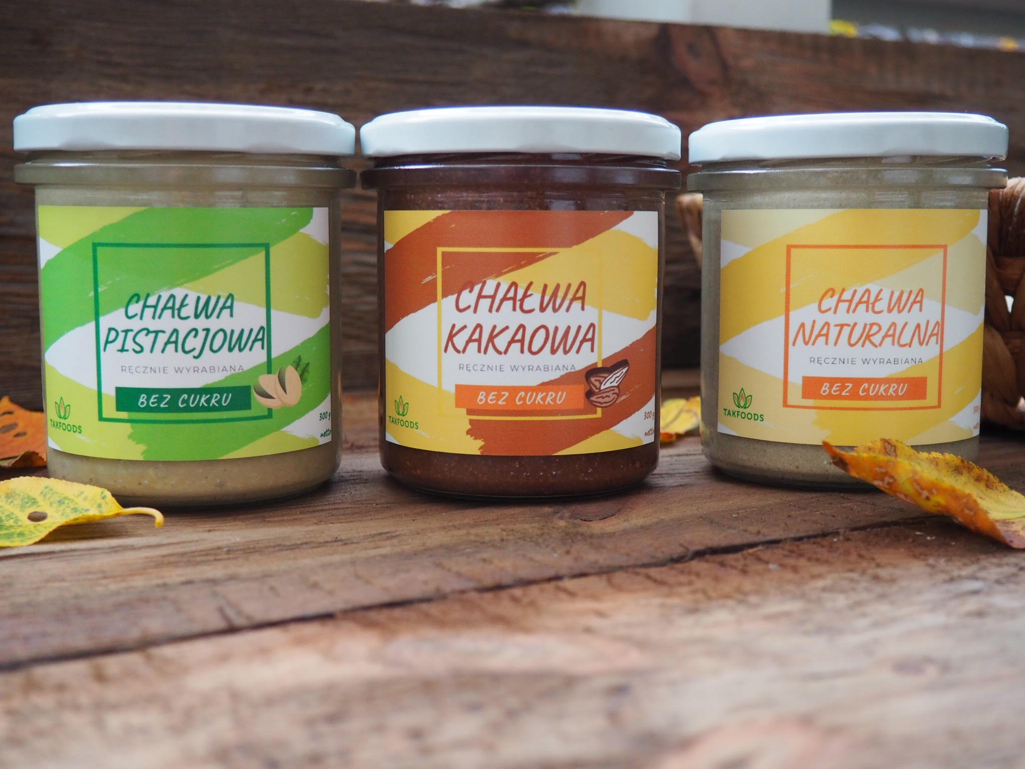 Trzy słoiki chałwy bez cukru: pistacjowa, kakaowa i naturalna, ustawione na drewnianym stole z jesiennymi liśćmi, prezentacja marki TAKFOODS.