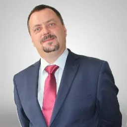 Portret mężczyzny w ciemnym garniturze i czerwonym krawacie, lekko pochylony w stronę kamery, na jednolitym szarym tle.