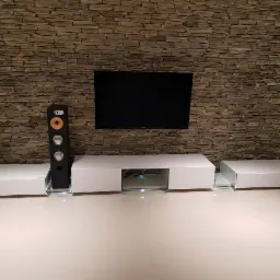 Biała szafka RTV z elementami szklanymi i głośnikiem na tle ściany z kamienia dekoracyjnego, pod wiszącym telewizorem.