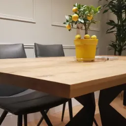 Stół z blatem z jasnego drewna na czarnych, geometrycznych nogach, otoczony szarymi krzesłami, z doniczką z cytrynami i zieloną rośliną w tle.