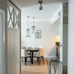 Jasne wnętrze z okrągłym stołem, krzesłami w odcieniach beżu, wiszącymi lampami ze szklanymi kloszami i dekoracyjną szafką z geometrycznym wzorem na drzwiach.