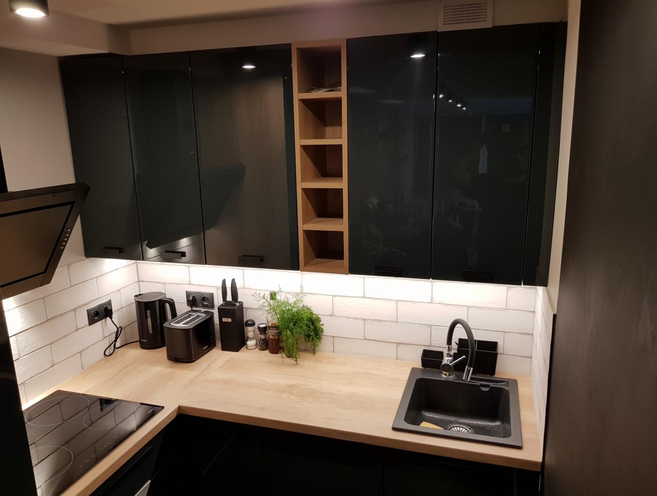 Nowoczesna kuchnia z ciemnymi, lakierowanymi szafkami górnymi, blatem w odcieniach drewna i białymi płytkami ściennymi imitującymi cegłę, oświetlona listwą LED pod szafkami.