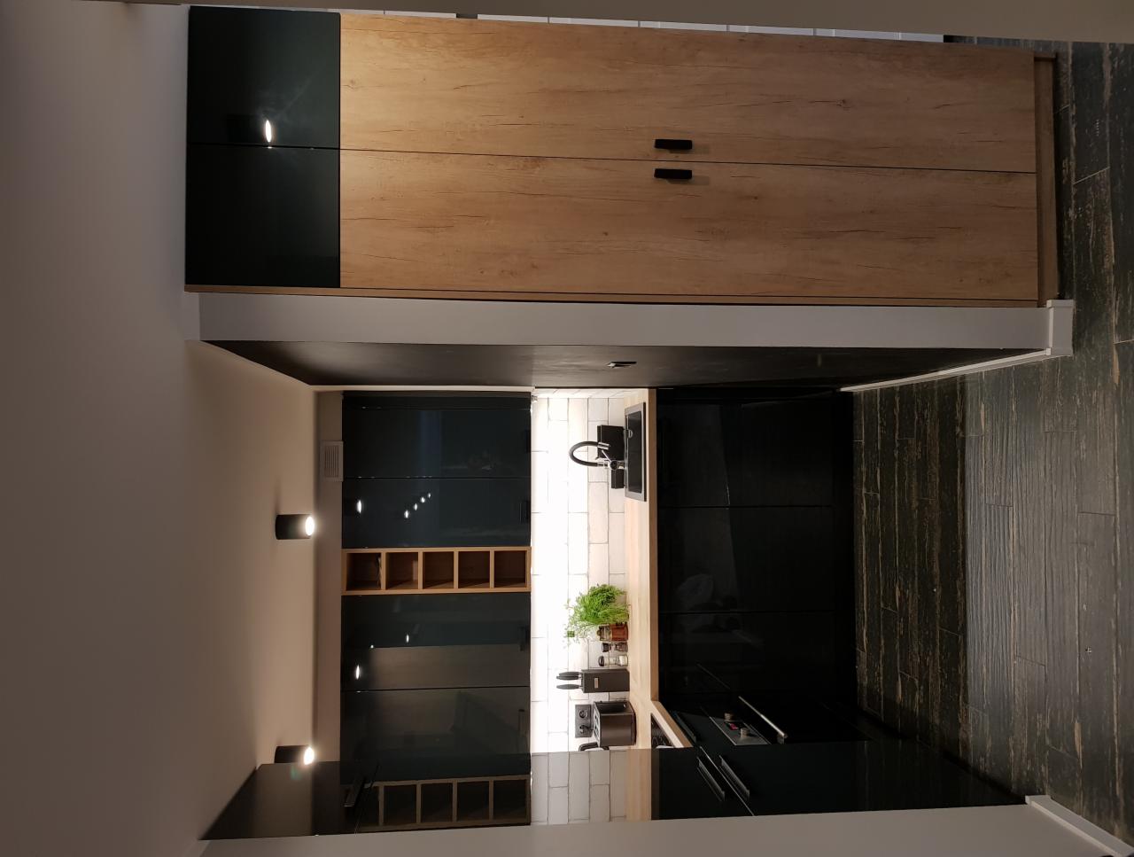 Nowoczesna kuchnia z czarnymi, lakierowanymi frontami szafek dolnych i drewnianymi szafkami górnymi, oświetlona punktowymi lampami ściennymi i listwą LED pod szafkami, z widocznym fragmentem blatu...