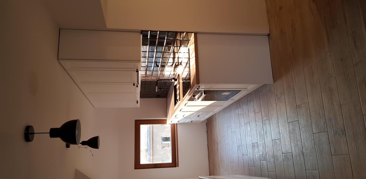 Nowoczesna kuchnia z białymi szafkami, drewnianym blatem, szarą ścianą nad blatem i drewnianą podłogą, oświetlona światłem dziennym z okna, z widoczną lampą na ścianie.