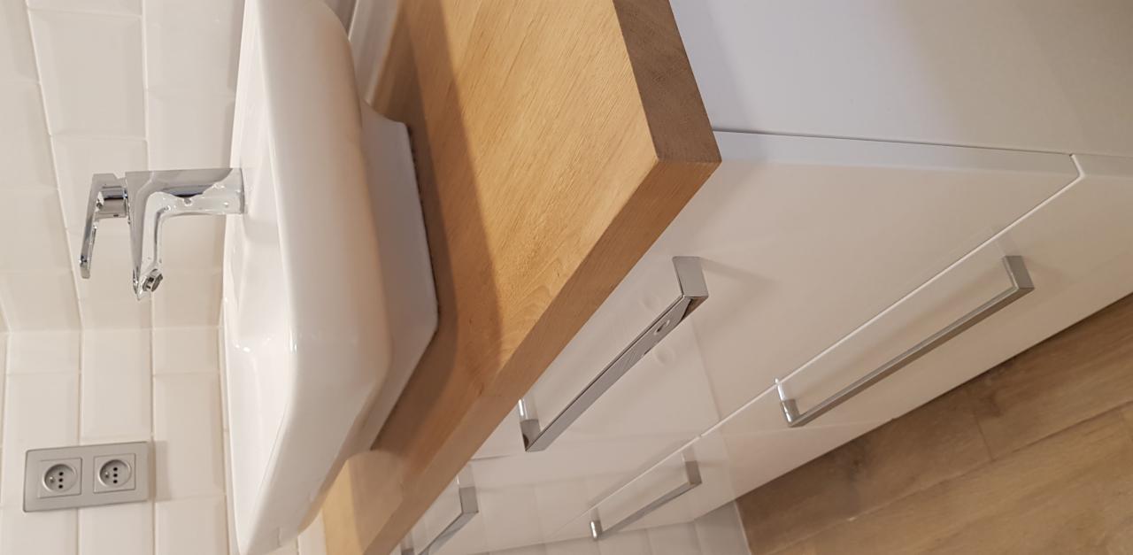 Nowoczesna łazienka z umywalką nablatową, drewnianym blatem i białymi szafkami z chromowanymi uchwytami, widok z dołu, białe płytki na ścianie i drewniana podłoga.