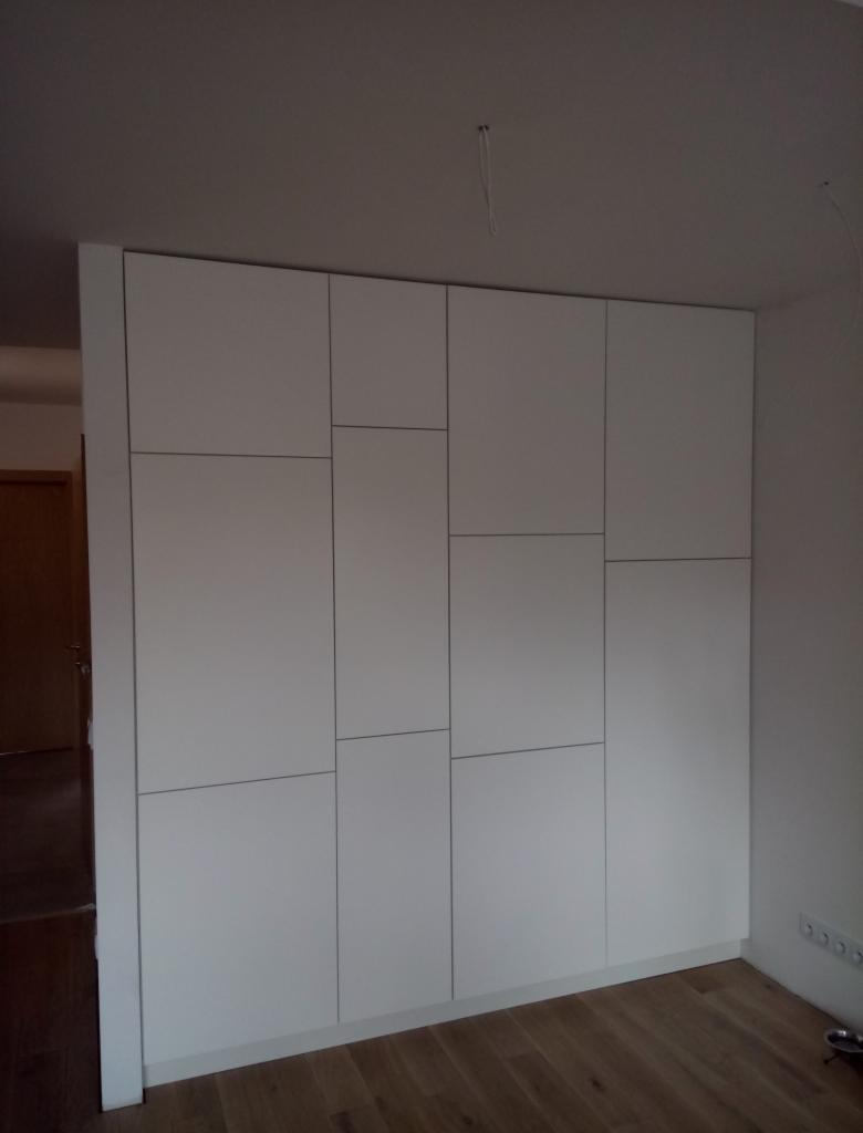 Biała szafa wnękowa z panelami o zróżnicowanych rozmiarach, widoczne gniazdka elektryczne na ścianie obok, podłoga z paneli drewnianych.