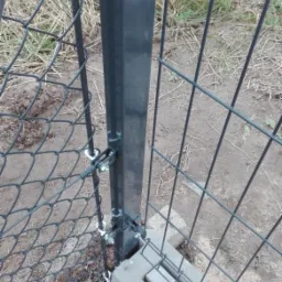 Fragment nowo montowanego ogrodzenia: czarny słupek przytwierdzony do podłoża betonowymi elementami, do słupka przymocowana siatka i panele ogrodzeniowe za pomocą śrub.