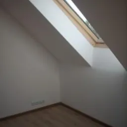 Poddasze ze świeżo pomalowanymi na biało ścianami, drewnianą podłogą z paneli i oknem dachowym z drewnianą ramą.