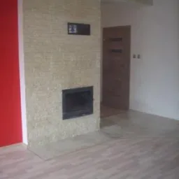 Wnętrze pokoju z kominkiem obłożonym kamieniem dekoracyjnym, drewnianą podłogą panelową i czerwoną ścianą po lewej stronie, widok na drzwi w tle.