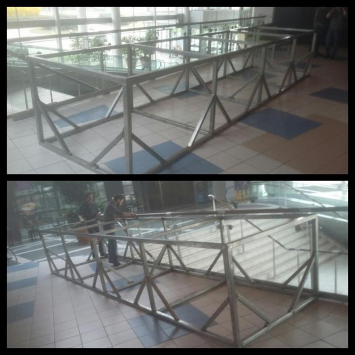 Metalowa konstrukcja nośna, prawdopodobnie pod przeszkloną podłogę lub kładkę, z widocznymi spawami i elementami mocującymi, ustawiona wewnątrz budynku z przeszkloną ścianą.