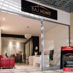Wnętrze biura nieruchomości Baj Home z widocznym logo nad wejściem, eleganckim żyrandolem i czerwonym fotelem, z tablicą reklamową przed wejściem.