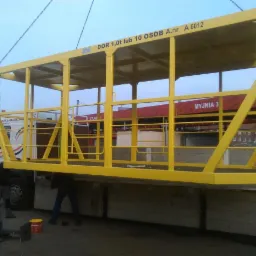 Żółta, metalowa platforma robocza, zawieszona na łańcuchach, transportowana na naczepie ciężarówki. Widoczni pracownicy przy ziemi i napis 'DOR 1,0t lub 10 OSÓB A.nr A 6012' na platformie.
