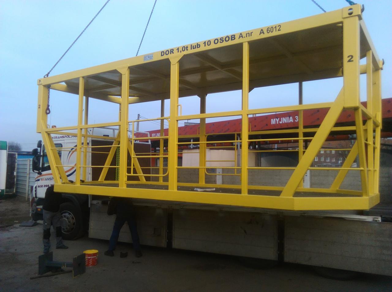 Żółta, metalowa platforma robocza, zawieszona na łańcuchach, transportowana na naczepie ciężarówki. Widoczni pracownicy przy ziemi i napis 'DOR 1,0t lub 10 OSÓB A.nr A 6012' na platformie.