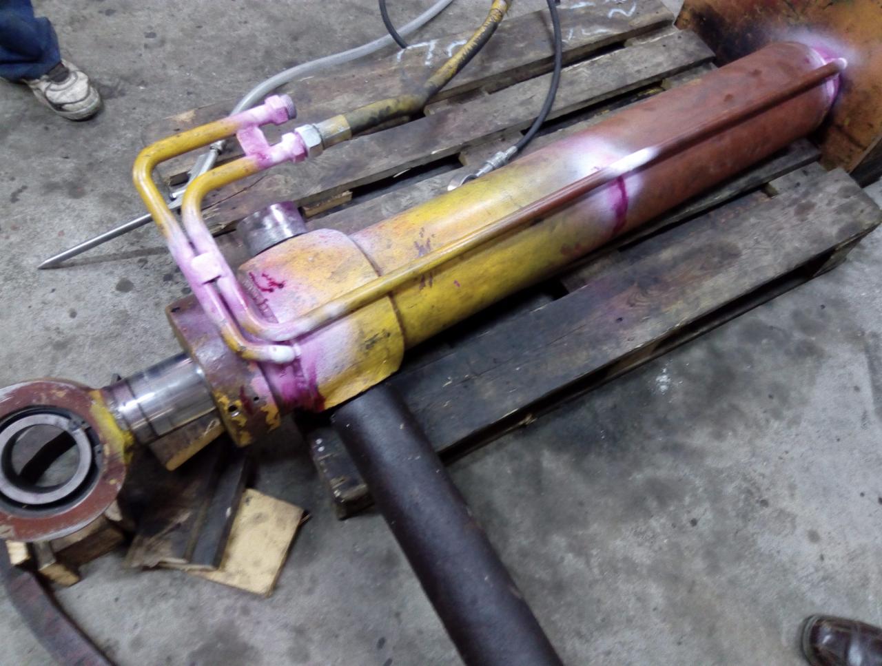 Hydrauliczny siłownik po teście szczelności barwnikiem penetracyjnym, leżący na drewnianej palecie. Widoczne liczne żółte przewody i złącza, a także ślady różowego barwnika.